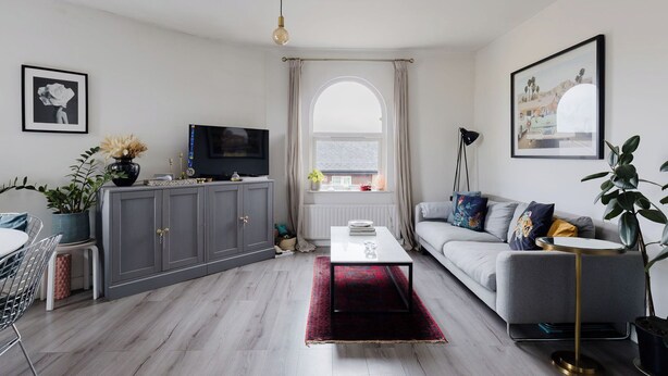 Un salon moderne avec un canapé gris, un téléviseur, un coin repas comprenant une table et des chaises, et une grande fenêtre avec des rideaux.
