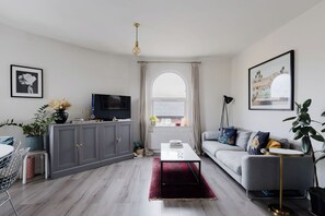Una sala moderna con un sofá gris, un televisor, un comedor con mesa y sillas, y un ventanal con cortinas.