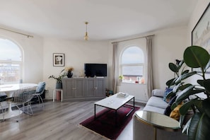 A modern living room with a dining area, grey cabinets, a flat-screen TV, and large windows.