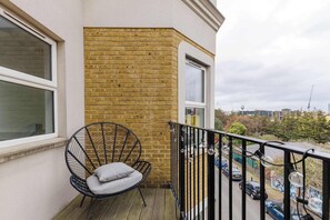 Een balkon met een zwarte metalen stoel en een kussen, een bakstenen muur, en uitzicht op een parkeerplaats met auto's en bomen.