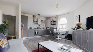 Une cuisine moderne avec des meubles gris, un coin repas avec une table et des chaises blanches, et un salon avec un canapé et une table basse.