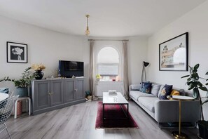 Een moderne woonkamer met een grijze bank, een salontafel, een televisie en een groot raam met gordijnen.