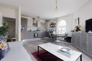 Een moderne keuken met grijze kastjes, een eetgedeelte met een witte tafel, en een woonkamer met een grijze bank.