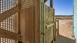 A wooden structure with a lattice design, a metal door, and a view of a sandy area with a fence and trees in the distance.