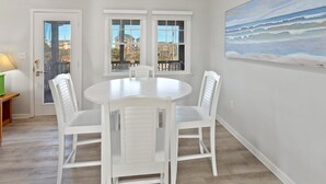 Un comedor con una mesa redonda y cuatro sillas blancas, ventanas amplias con vista a edificios, y un cuadro de una playa en la pared.