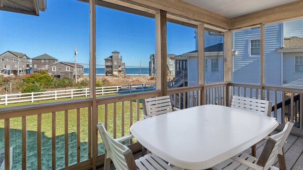 Una terraza cubierta al aire libre con una mesa y sillas blancas, con vistas a una playa con casas y una torre de salvavidas.