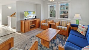 Una sala de estar con televisión, muebles de madera, un sofá azul y vistas a casas a través de las ventanas.