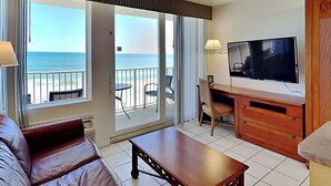 Ein Hotelzimmer mit einer großen Schiebeglasdoor zum Balkon mit Blick auf das Meer. Es gibt einen Holztisch mit Stuhl, einen an der Wand montierten Fernseher und ein Ledersofa.