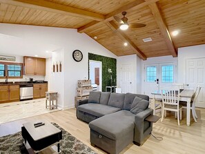 A modern living room with a sectional sofa, a dining table with chairs, a kitchen area, and a ceiling fan.