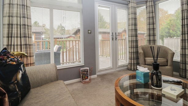 Ein Wohnzimmer mit einer Couch, einem Couchtisch und einem Sessel. Zwei Fenster mit Jalousien, eine Tür und ein Blick auf einen Garten.
