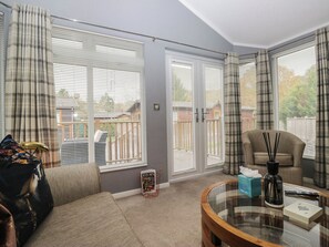 A living room with a sofa, a coffee table, and a chair. There are two windows with blinds, a door, and a view of a backyard. - 32 Grasmere (Windermere)