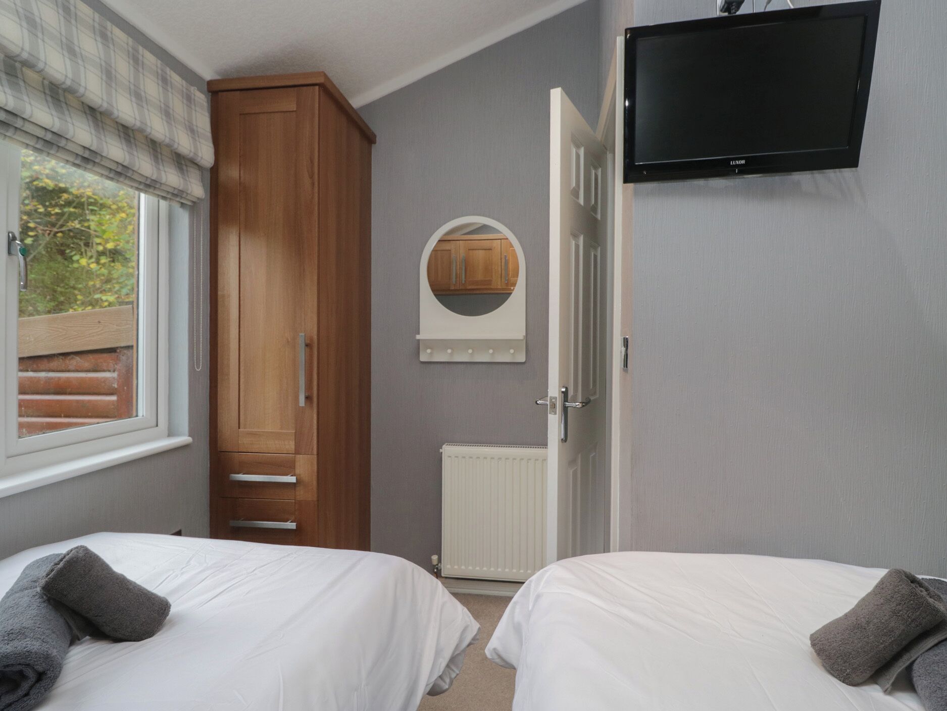 A bedroom with two beds, a wardrobe, a television, and a window with a view of greenery.