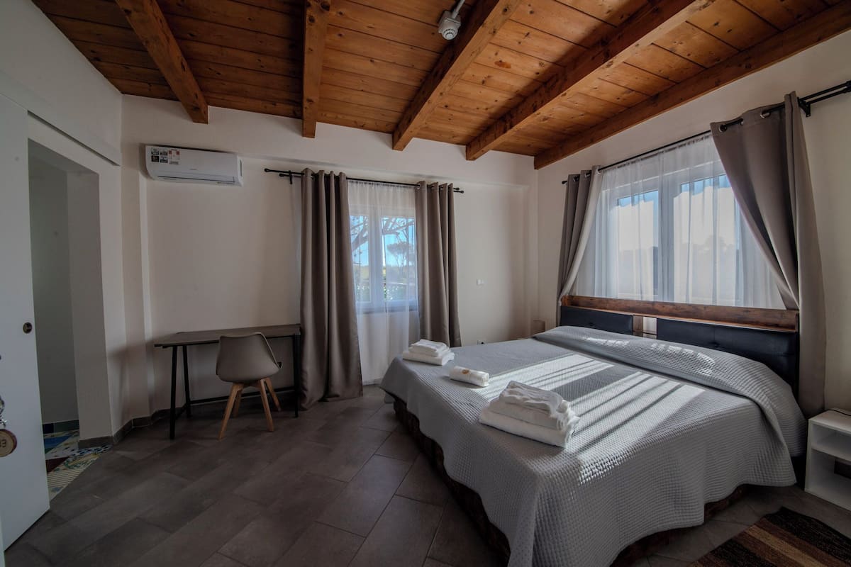 A bedroom with a large bed, a desk, and a chair. There is an air conditioning unit on the wall and a window with curtains.