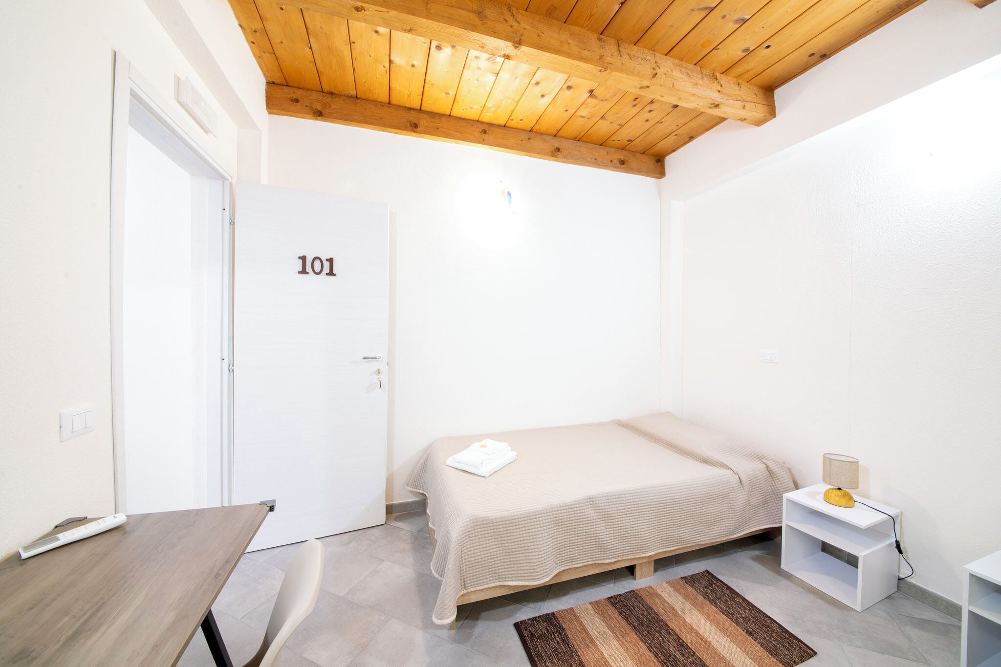 Una camera da letto con un letto, una scrivania, una sedia, un tavolino, un comodino e una porta con la scritta '101'.