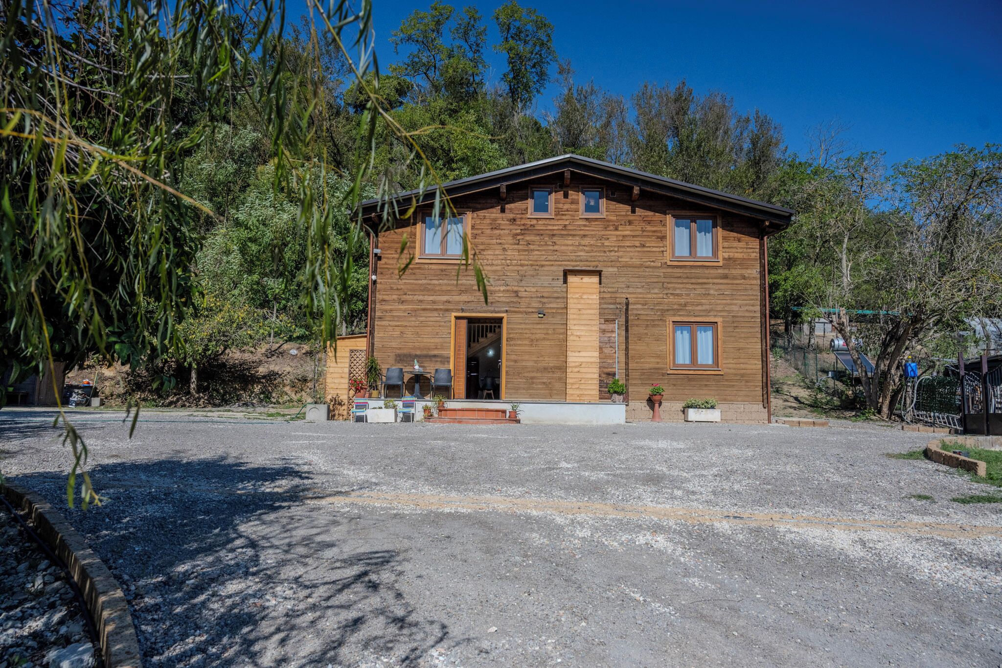 A two-story wooden house with a front porch, a small outdoor seating area, and a gravel driveway.