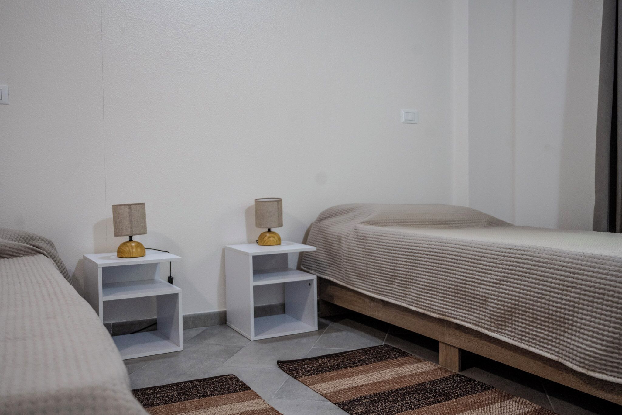 A small, clean room with two beds, a nightstand with lamps, and a patterned rug.