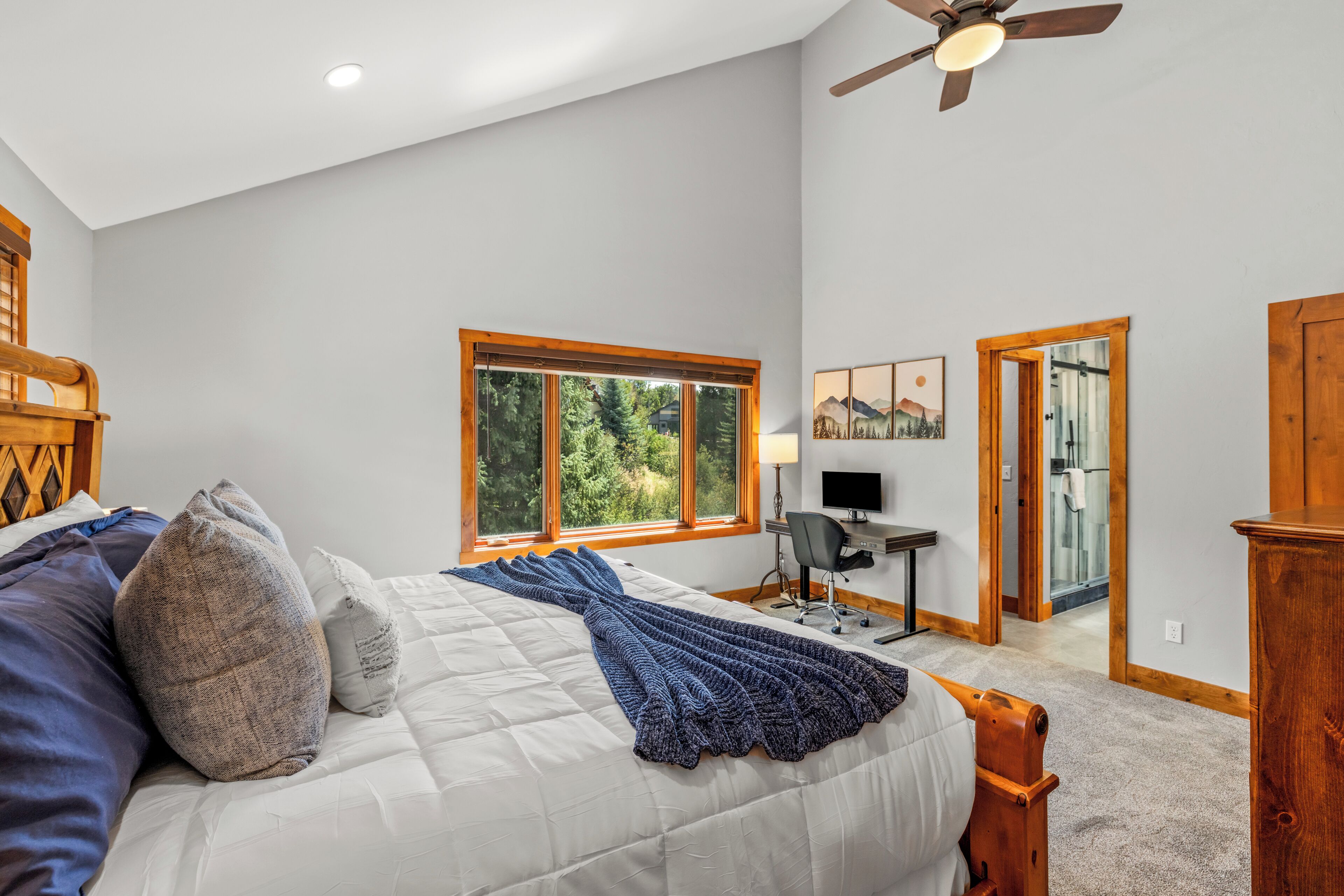 A bedroom with a bed, a desk, a chair, a ceiling fan, and a window with a view of trees.