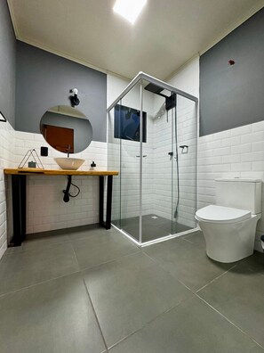 A modern bathroom with a glass-enclosed shower, a white toilet, and a wooden vanity with a round mirror. - Lumira House - Bombinhas - SC (Bombinhas)