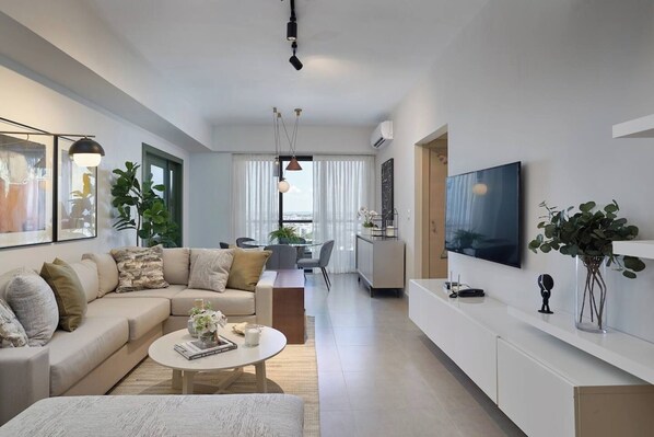A modern living room with a sofa, coffee table, and TV. There is a dining area with a table and chairs.