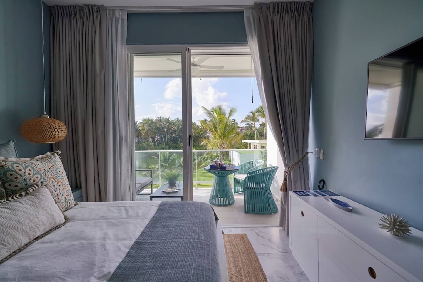 Ein Schlafzimmer mit Bett, Schreibtisch, Stuhl, einem Balkon mit Tisch und Stühlen sowie Blick auf Palmen.