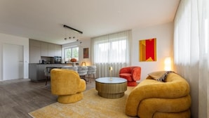A modern living room with a sofa, armchair, and coffee table. There is a kitchen in the background with a dining area. - Lorenzhaus Exclusive Residence: Deluxe apartment with 3 bedrooms (Sankt Lorenz)