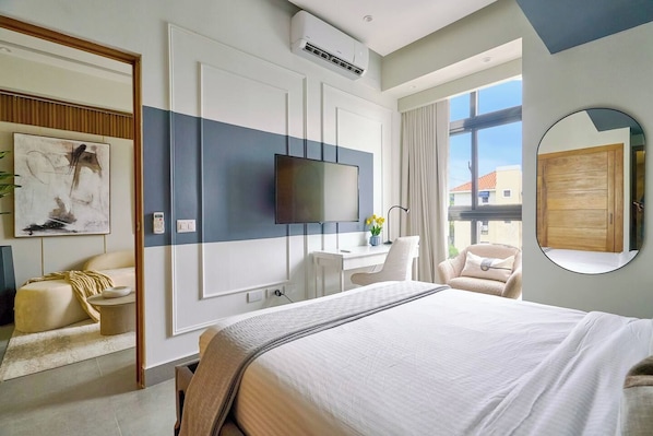 A hotel room with a large bed, a desk with a chair, a television, and a window with a view of buildings.