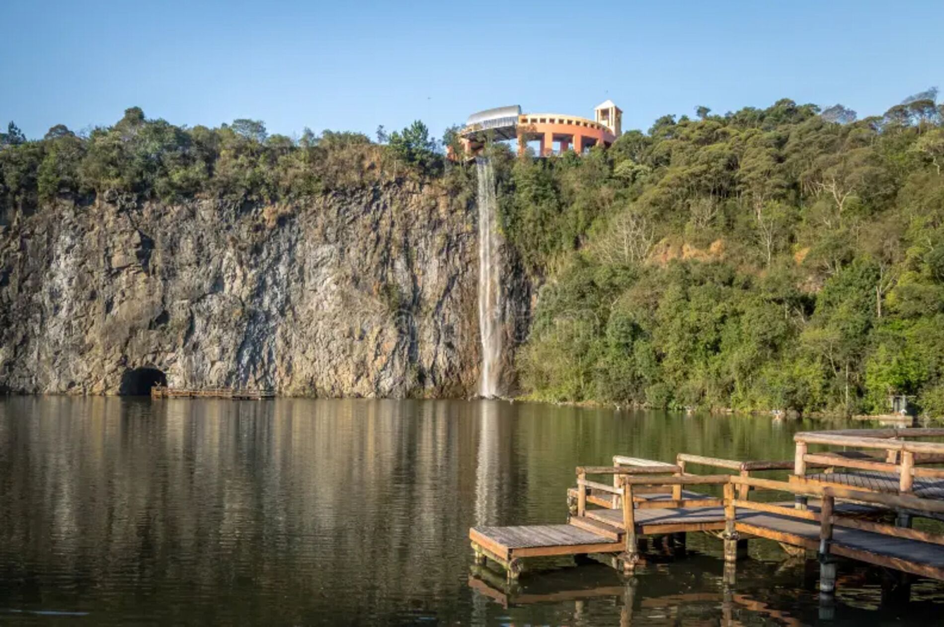 Una cascada se precipita en un lago, rodeada por un acantilado rocoso y abundante vegetación. Un muelle de madera se adentra en el agua, y se observa un edificio con estructura de cúpula en el acantilado.