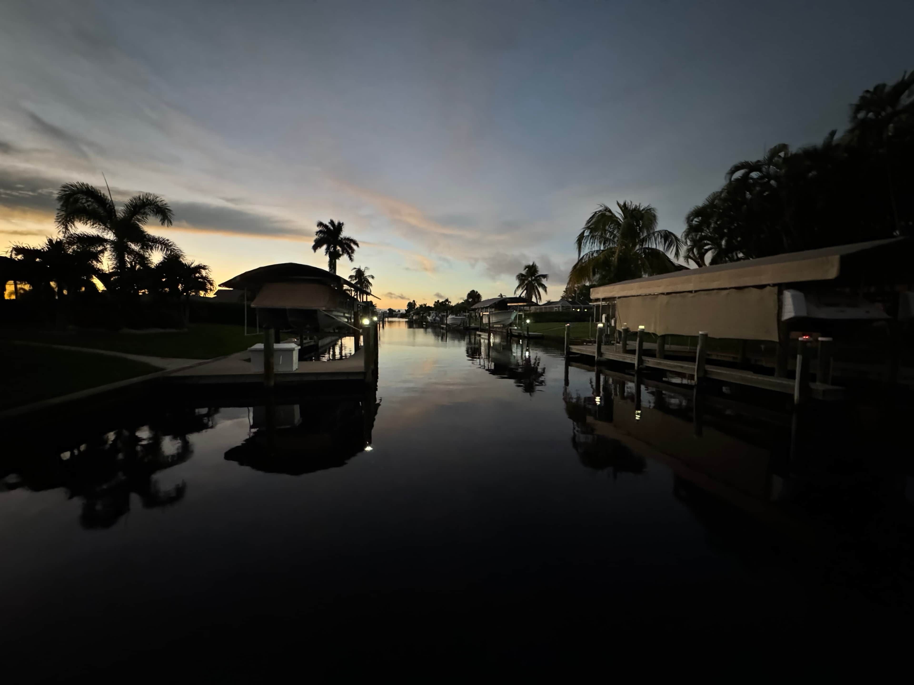 A calm water body with a dock and a covered seating area, surrounded by palm trees and a clear sky.