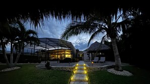 A modern house with a glass facade, surrounded by a well-maintained lawn and palm trees, leading to a thatched-roof structure. - Villa Palma Encantada Gulf Access & Pet Friendly (Cape Coral)
