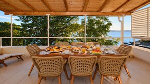 Un espace extérieur abrité, aménagé pour prendre les repas, avec des chaises en osier et une table dressée pour le petit-déjeuner, offrant une vue sur un plan d’eau et des arbres.