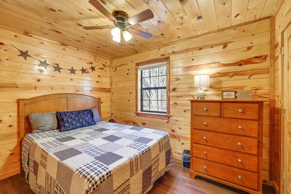 A wooden cabin bedroom with a bed, dresser, and ceiling fan. - Riverfront Fishing Cabin | 3BR Sleeps 6 | Great Weekday Rates (Broken Bow)