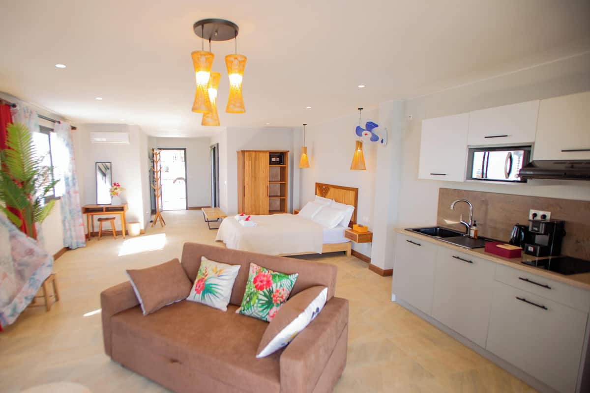 A modern hotel room with a bed, a sofa, a kitchen area, and a dining area.