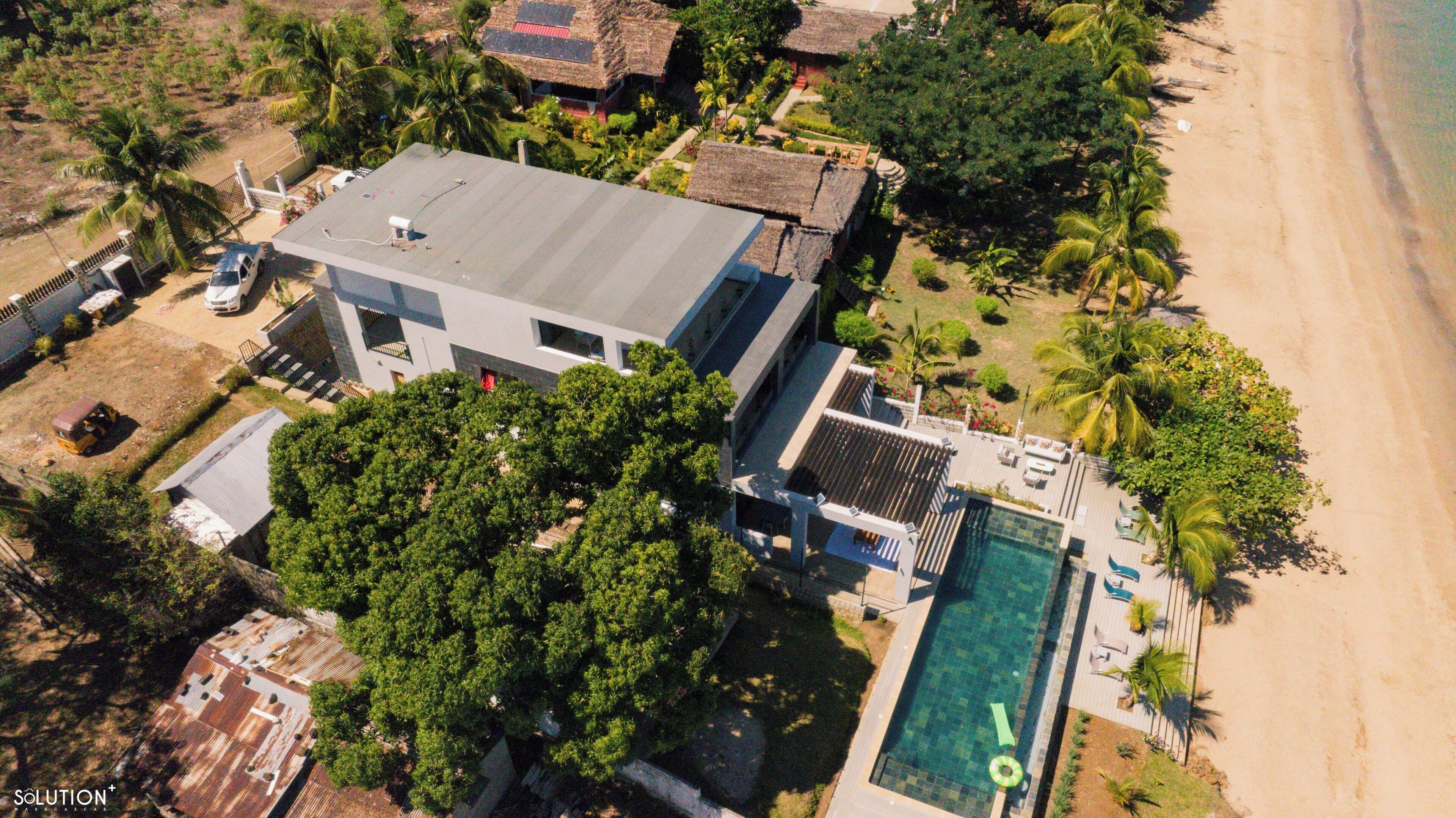 Vista aérea de una casa moderna con piscina, rodeada de palmeras y una playa.