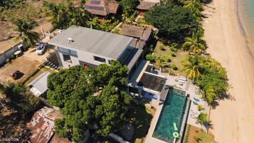 Vista aérea de una casa moderna con piscina, rodeada de palmeras y una playa.