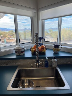 A kitchen with a stainless steel sink, a window with a view of a landscape, and kitchen items like a pot, a fruit basket, and a pitcher.