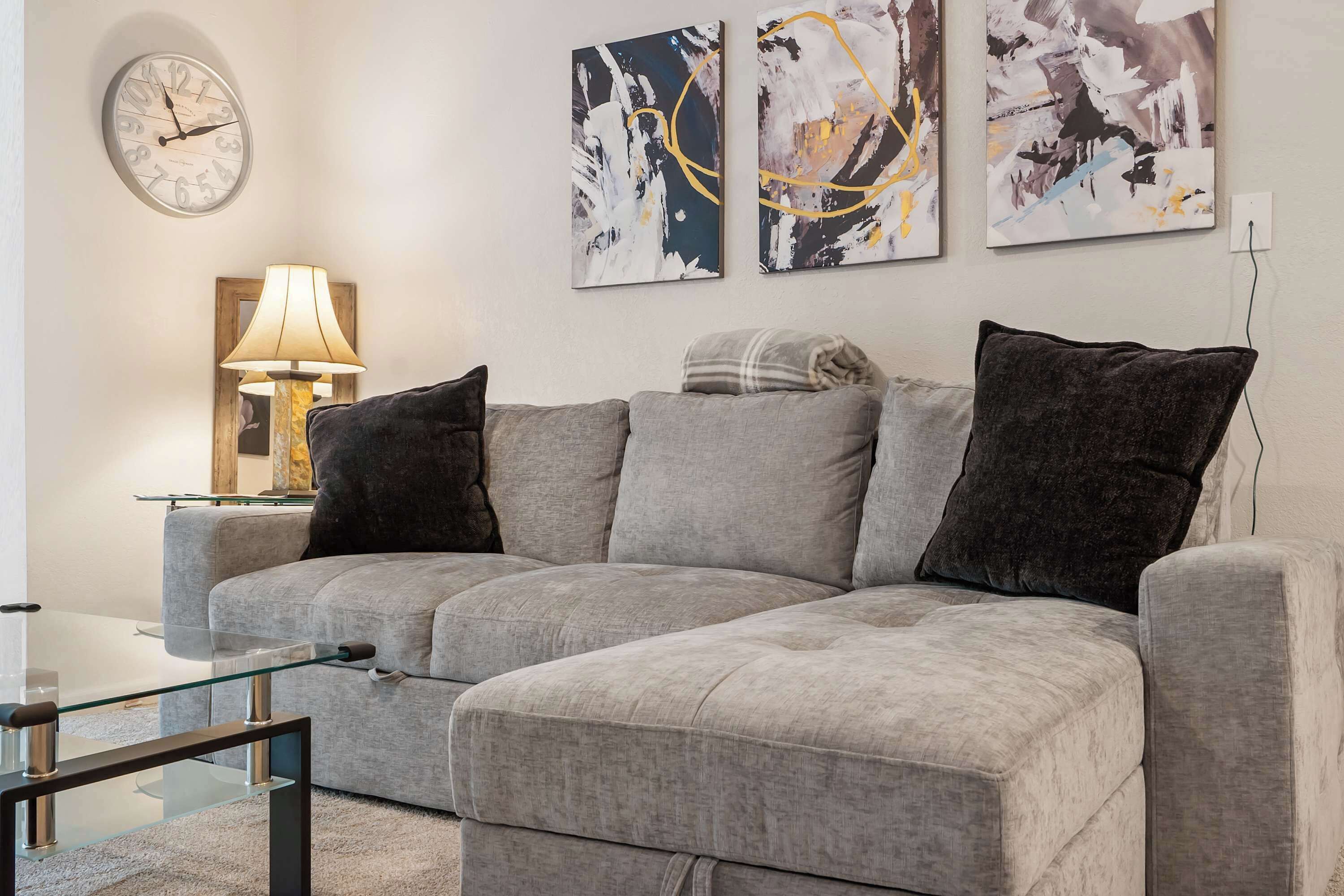 A modern living room with a grey sectional sofa, a glass coffee table, a wall clock, abstract paintings, and a floor lamp.