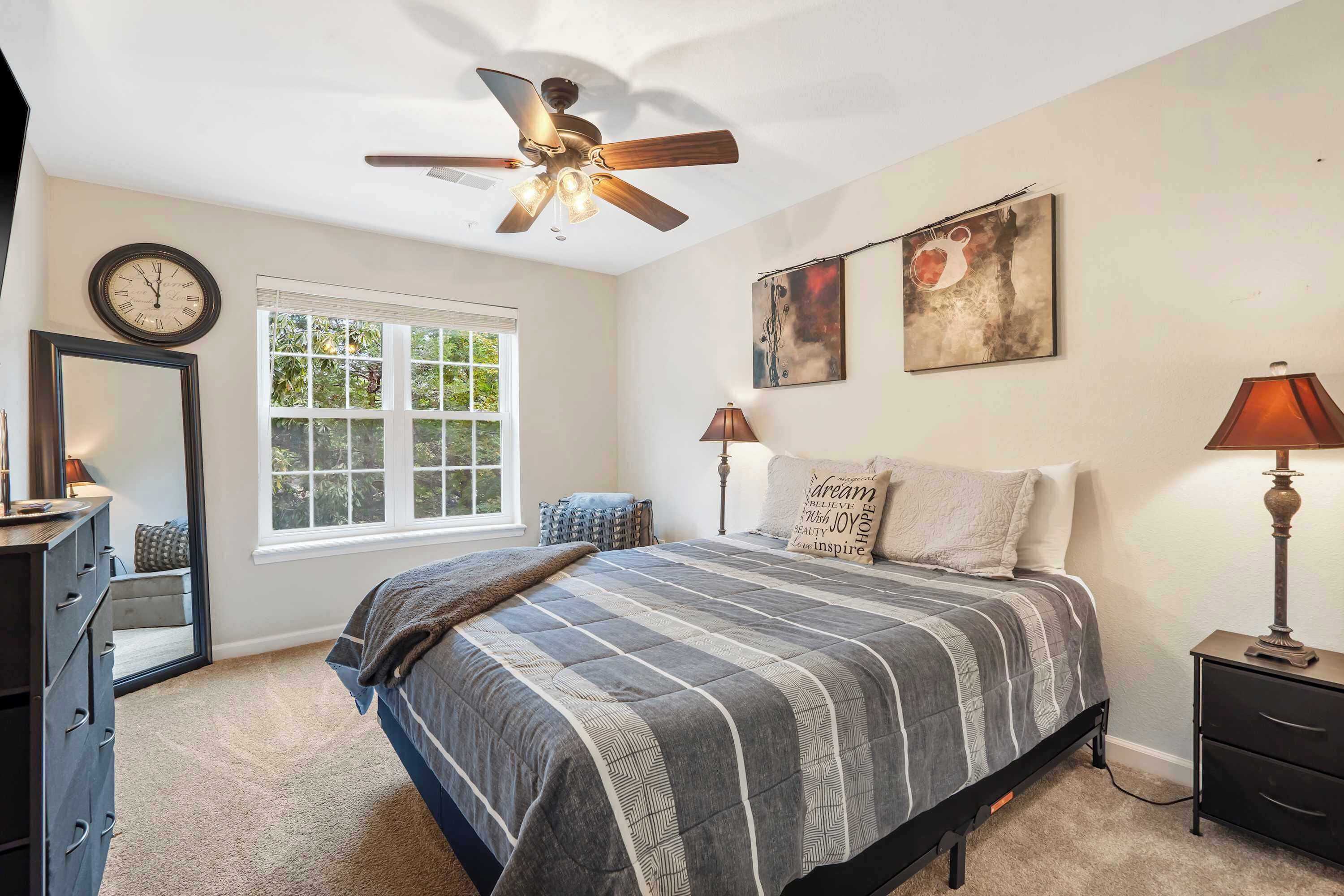 A bedroom with a bed, a ceiling fan, a window, a dresser, and two wall-mounted lamps.