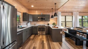 A modern kitchen with stainless steel appliances, wooden flooring, and a central island with seating. - Modern Smoky Mountain Cabin Near Dollywood – Indoor Pool, Hot Tub & Arcade Fun (Pigeon Forge)