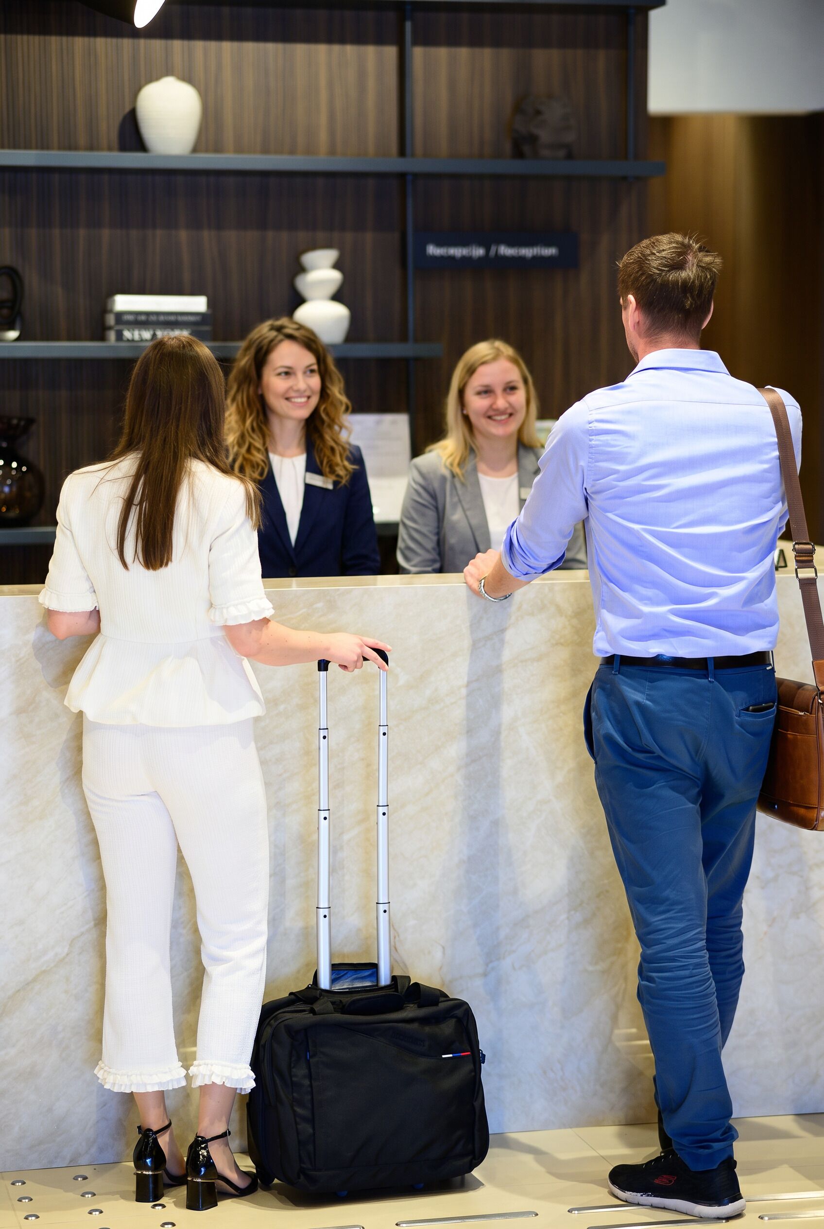 A reception area with a marble counter, a black luggage trolley, and a person with a brown shoulder bag.