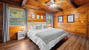 Un dormitorio de cabaña de madera con cama, mesitas de noche, ventilador de techo y cuadros en las paredes.