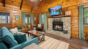 Interior acogedor de una cabaña de madera con chimenea de piedra, un televisor montado encima y un espacio de estar cómodo con sofá y sillones.