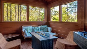 Una terraza de madera con mobiliario de mimbre, una chimenea y amplios ventanales con vistas a un bosque.