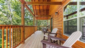 A wooden deck with Adirondack chairs, a table, and a window overlooking a forest.