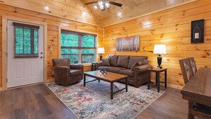 A cozy cabin interior with a wooden ceiling, a ceiling fan, a large window, a brown leather sofa, a coffee table, and a rug.