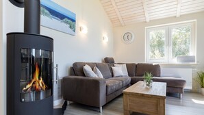 A modern living room with a wood-burning stove, a brown sofa, a wooden coffee table, and a large window with a view of greenery. - Ferienhaus in Dagebuell im skandinavischen Stil (Dagebüll)