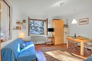 A cozy living room with a blue sofa, a dining table with chairs, a TV, and a window with a snowy view. - Appartamento Chez Mavi Cond Il (Sauze d'Oulx)