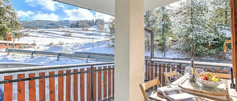 A balcony with a view of a snow-covered landscape, a wooden table with a bowl of fruit, and a bicycle.