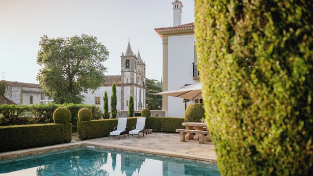 Uma piscina com espreguiçadeiras e um banco, rodeada por sebes bem aparadas e um edifício com uma torre de sino ao fundo.