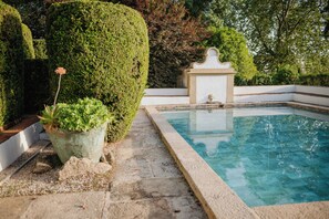 Ein Pool mit Steingrenze, umgeben von ordentlich geschnittenen Hecken und einer kleinen weißen Struktur.