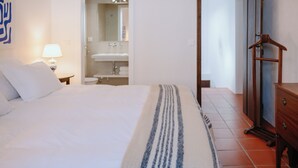 Quarto com uma cama, uma mesa de cabeceira com abajur, cômoda de madeira e um banheiro visível por uma porta aberta.